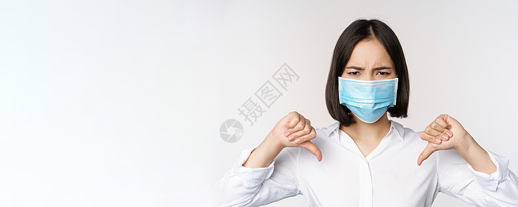 在医疗面罩上假面具的亚洲妇女脸部肖像显示拇指低下 表情失望 疲倦 站立在白色背景之上冒充管理人员办公室成人售货员员工商务女孩面具图片