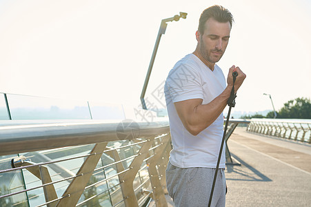 英俊的欧洲年轻运动员 在户外锻炼时使用橡胶阻力带的运动员 黎明时分 中年运动员在玻璃城桥上使用弹性扩张器锻炼二头肌图片