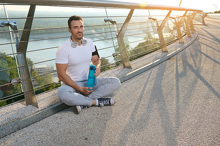 帅气的白人肌肉男 坚定的运动员 戴耳机的运动员在黎明户外锻炼后放松 拿着水瓶 夏天坐在玻璃城桥上图片