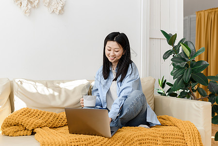 年轻的亚洲女性坐在带格子花呢沙发上 喝着热茶或咖啡 看连续剧或电影 或者在社交媒体上放松身心图片