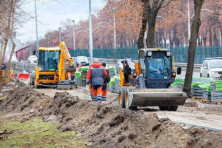 紧凑型平路机沿着正在施工的人行道行驶 前方是施工现场 一队道路工人模糊不清图片