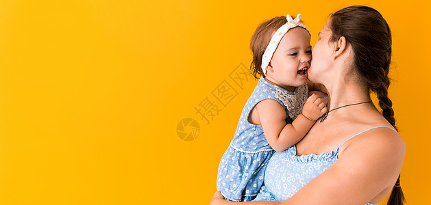 母亲 炎热的夏天  身穿蓝色裙子的年轻怀孕快乐微笑的母亲 抱着小女儿蹒跚学步的兄弟姐妹 看着黄色背景的矮树丛空间上的眼睛亲吻感情图片
