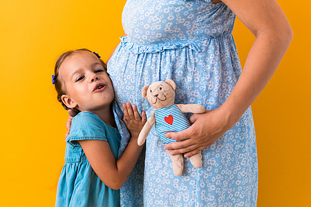 母亲 爱情 童年 怀孕 炎热的夏天  裁剪肖像怀孕的无法辨认的母亲蓝色连衣裙小学龄前女儿女孩兄弟姐妹泰迪熊拥抱黄色背景的妈妈肚子图片