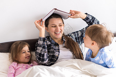 幸福的家庭 特写慈爱的母亲和女儿儿子躺在床上 两个孩子睡衣在床上 孩子们读有趣的故事书 学龄前微笑的小女孩在睡觉前享受童话故事图片