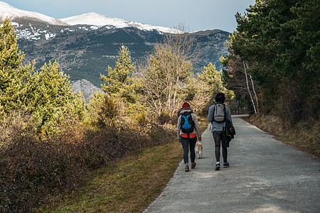 年轻夫妇背着背包和一条狗在路上 背景是雪山 男人和女人在阳光明媚的日子徒步旅行图片