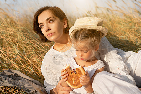 母亲和女儿在野餐时坐在高黄草地上吃面包父母稻草两个人孩子女性包子家庭衣服农村草帽图片