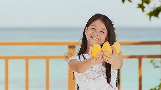 一个穿着白色裙子的迷人的菲利平女学生 长发正装着芒果 在她手中 阳光 蓝海 童年 娱乐 笑声喜悦微笑享受孩子食物橙子公园水果小吃图片