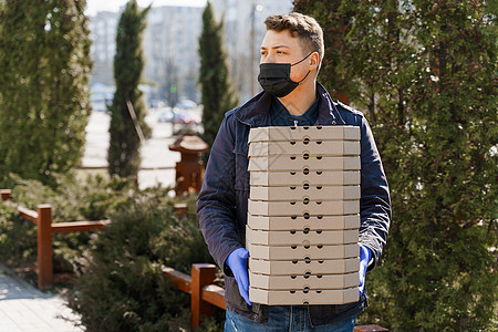 在送餐公司为年轻人工作 戴着黑色医用口罩和蓝色无菌手套的快递员 手里拿着很多披萨盒图片