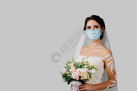 在Corona病毒(covid-19)隔离期穿戴医疗面具和婚礼花束的新娘 有吸引力的女孩在白色背景的工作室里用摄像头看图片