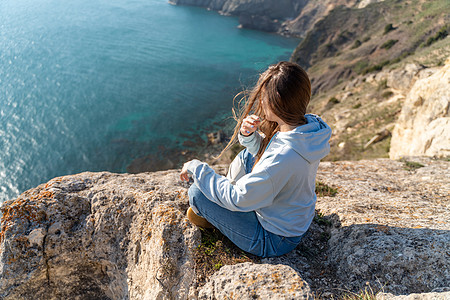 女性游客享受海山地貌的日落 坐在海面上的岩石上户外 穿着牛仔裤和蓝色连帽衫 健康生活方式 和谐与冥想 续山脉瑜珈阳光手势杯子沉思图片