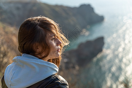 女性游客享受海山地貌的日落 在海面上的一个岩石上户外露宿 穿着牛仔裤 蓝连帽衫和黑皮夹克 健康生活方式 和谐与默思成人假期天空自图片