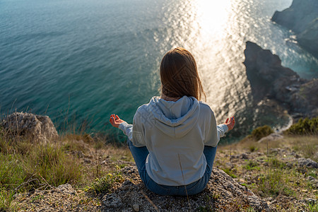 女性游客享受海山地貌的日落 坐在海面上的岩石上户外 穿着牛仔裤和蓝色连帽衫 健康生活方式 和谐与冥想 续假期瑜伽山脉喜悦海洋太阳图片