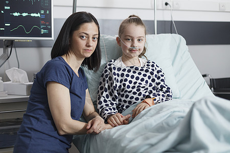 年轻担心的母亲坐在儿科病房里微笑着生病的小女儿旁边 看着相机图片