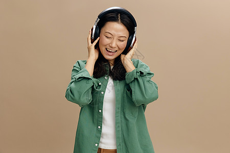 放松享受最喜欢的歌曲可爱的亚洲学生年轻女子在卡其绿色衬衫触摸耳机在米色柔和的工作室背景上孤立地摆姿势 酷时尚优惠 音乐应用平台广图片