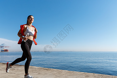 微笑的女运动员沿着海边奔跑并欣赏美景的户外拍摄 在海边慢跑的女运动员码头有氧运动女孩行动赛跑者锻炼跑步海洋运动训练图片