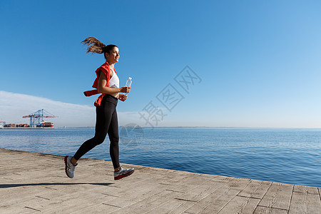 运动女青年在海边赛车场上跑动的侧视角 体育女运动员用瓶水在海洋附近慢跑活动女孩成人天空赛跑者训练行动锻炼慢跑者阳光图片