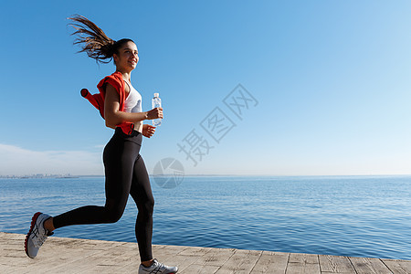 在海边赛道上跑动的有吸引力和健康的健身女子的侧面景色 女孩在早晨慢跑 微笑成人阳光锻炼码头训练慢跑者天空运动女性有氧运动图片