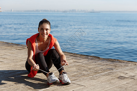 坐在码头的有吸引力 微笑着笑容的女运动员在外门拍摄 并在锻炼和海上训练期间系鞋带行动有氧运动福利运动女孩阳光赛跑者跑步海洋天空图片