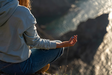 女性游客享受海山地貌的日落 坐在海面上的岩石上户外 穿着牛仔裤和蓝色连帽衫 健康生活方式 和谐与冥想 续喜悦成人太阳阳光自由手势图片