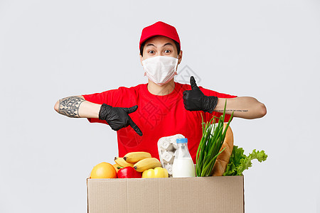 网上购物 食品配送和冠状病毒流行病概念 身穿红色制服 医用口罩和手套的超凡魅力送货员 指着杂货包裹竖起大拇指并表示认可购物载体命图片