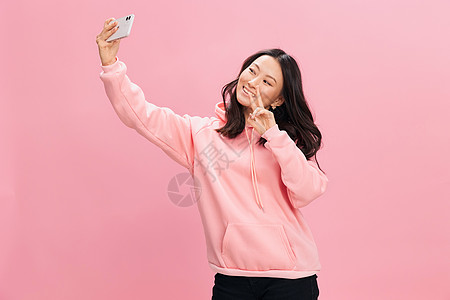 手机通话身穿粉红色连帽运动衫 面带微笑的亚洲年轻女士在粉红色工作室背景下孤立地进行自拍视频通话远程通信 好报价 小工具成瘾社交媒体概念互背景