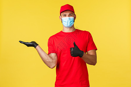 大流行期间的快递 covid19 安全运输 网上购物概念 身着红色制服 医用口罩和手套的微笑快递员展示产品并竖起大拇指表示认可工图片