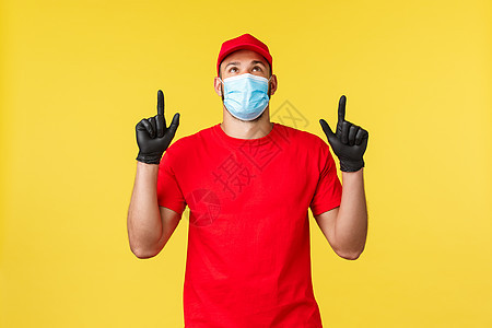 大流行期间的快递 covid19 安全运输 购物概念 穿着红色制服 医用面具和手套的满意微笑的快递员 指着并抬头看着宣传片 黄色图片
