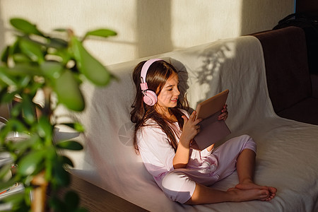 一个穿粉红色耳机的小可爱女孩坐在沙发上 看着数字平板电脑电影音乐游戏学习技术时间婴儿阳光互联网闲暇图片