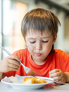 穿红色T恤的小孩用刀叉吃煎饼 长着面部令人迷惑的好奇男孩 早餐吃甜点图片