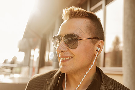 一位笑着微笑的欧洲青年商务人士在太阳镜和耳机上露面 听户外街边音乐时听到的正面景象列表闲暇手机衣服乐趣男人街道电话城市技术图片