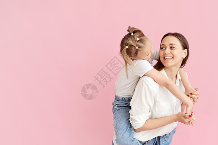 穿着浅色衣服的妈妈和可爱的小女孩玩得很开心 小女儿坐在母亲的背上 与柔和的粉红色背景隔离 工作室肖像图片