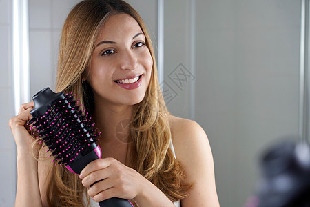可爱的女孩拿着圆梳子烘发机 在家中的洗手间打理头发 年轻女性使用沙龙一脚吹发机和挥发器图片