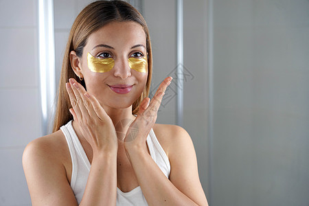 美丽的女孩在浴室里用金色抗衰老眼膜照镜子 皮肤护理女性在眼睛下方贴上织物面膜以减少眼袋图片