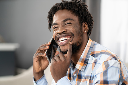 快乐微笑的年轻非洲裔美国男子手持智能手机通电话 看着别处 坐在沙发上进行当前对话的快乐男人的画像 社交媒体概念图片