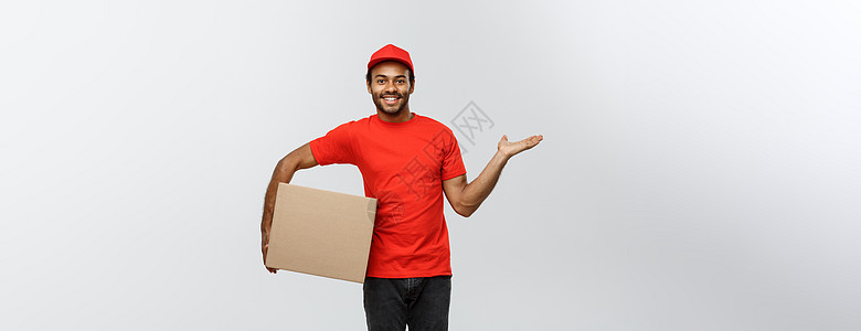 送货概念快乐的非洲裔美国送货员的肖像 指着手展示一个盒子包裹 隔绝在灰色演播室背景 复制空间男人工作手势男生职业工人相机船运男性图片