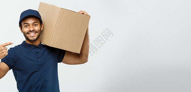 送货概念快乐的非洲裔美国送货员的肖像 指着手展示一个盒子包裹 隔绝在灰色演播室背景 复制空间服务工人男生导游相机男人白色工作室微图片