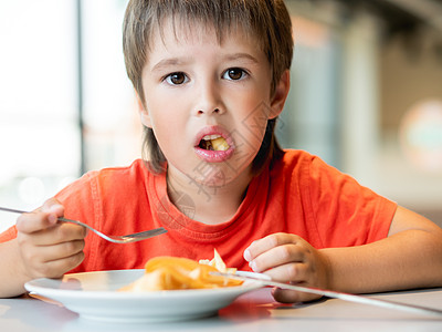 穿着红色T恤的小孩用刀和叉子吃食物 而脸部令人迷惑的快活男孩图片