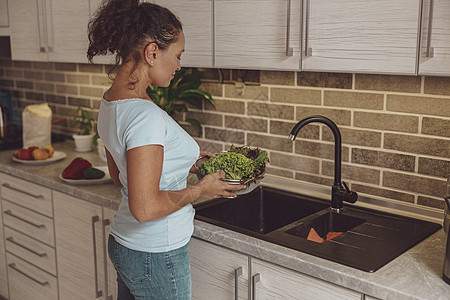 一个女人站在厨房水槽前 拿着一盘绿绿菜图片
