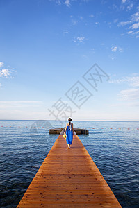 穿蓝裙子的女孩站在海边的木头码头上 有云的美丽的天空 度假概念 去欧洲旅行 (笑声)图片