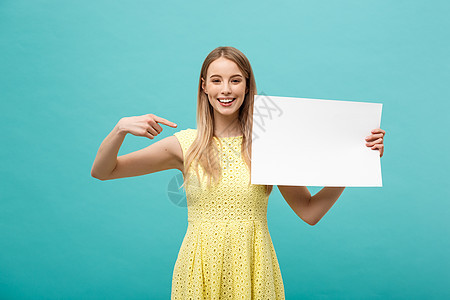 身着黄色裙子的年轻女子的肖像指向侧面白白空白板 孤立在蓝背景之上微笑蓝色黑发标语海报广告牌木板商业女士广告图片