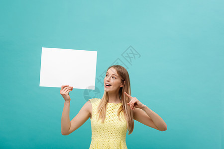 身着黄色裙子的年轻女子的肖像指向侧面白白空白板 孤立在蓝背景之上蓝色女士黑发学生商业微笑女孩成人广告牌广告图片