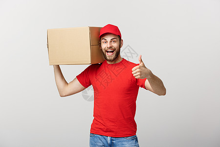 坐在灰色背景下站立时 拿着纸板盒举起大拇指的年轻英俊送货员拇指纸盒船运邮政服务工作职业邮递员工人成人图片