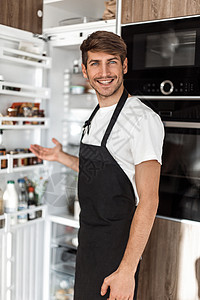 年轻人站在舒适的厨房里微笑生活冰箱福利衬衫食物美食围裙家庭男性图片