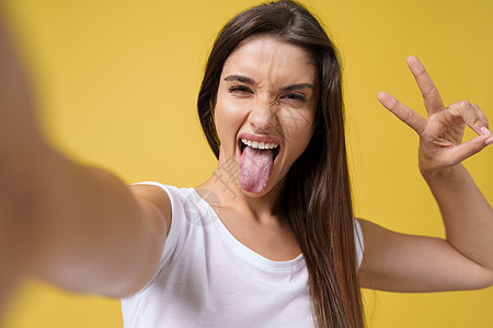 令人愉快的迷人女孩在工作室里自拍并大笑 留着棕色头发的漂亮年轻女子在明亮的黄色背景下自拍手机成人女士电话幸福微笑旅行照片快乐乐趣图片