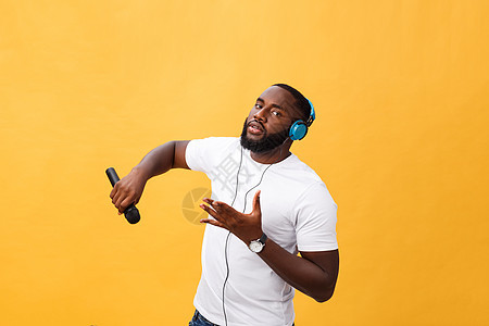 欢快积极 别致 英俊的非洲男子手持麦克风 头戴耳机听音乐唱歌 享受黄色背景中孤立的周末假期的肖像寒意青少年舞蹈男人乐趣微笑头发手图片