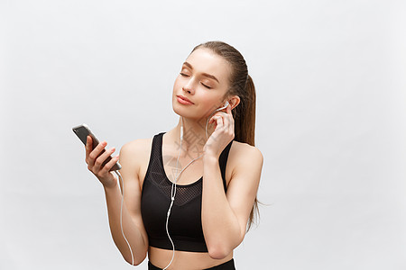 年轻女运动员的孤立镜头拥有健康的运动身体 戴着耳机听音乐 拿着智能手机 孤立在灰色的背景冒充音乐运动装训练女孩旋律享受电话工具锻图片