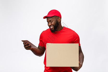 送货概念快乐的非洲裔美国人送货员的画像 穿着红布拿着盒子包裹 隔绝在灰色演播室背景 复制空间礼物红色工人相机手臂邮递员商业导游男图片