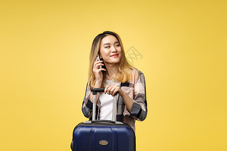 快乐的亚洲女性旅行者的画像 带着手提箱 在孤立的黄色背景下看着手机商业背包女孩商务电话人士行李白色牛仔裤女士图片