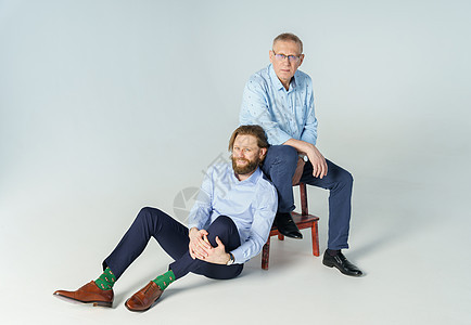 父亲坐在椅子上 儿子坐在白色背景上 两个男人都看着镜头 一个戴着屈光度眼镜的老人 一个留着胡须和长发的时尚年轻人男子两个人朋友们图片
