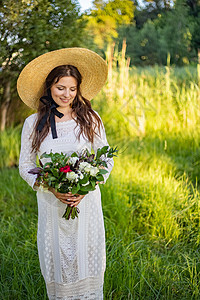 一位年轻漂亮的新娘化妆和发型穿着波西米亚风格的婚纱站在草地上 她手里拿着一束鲜花 肩上的大草帽夫妻女性蓝色男人庆典乡村裙子订婚婚图片
